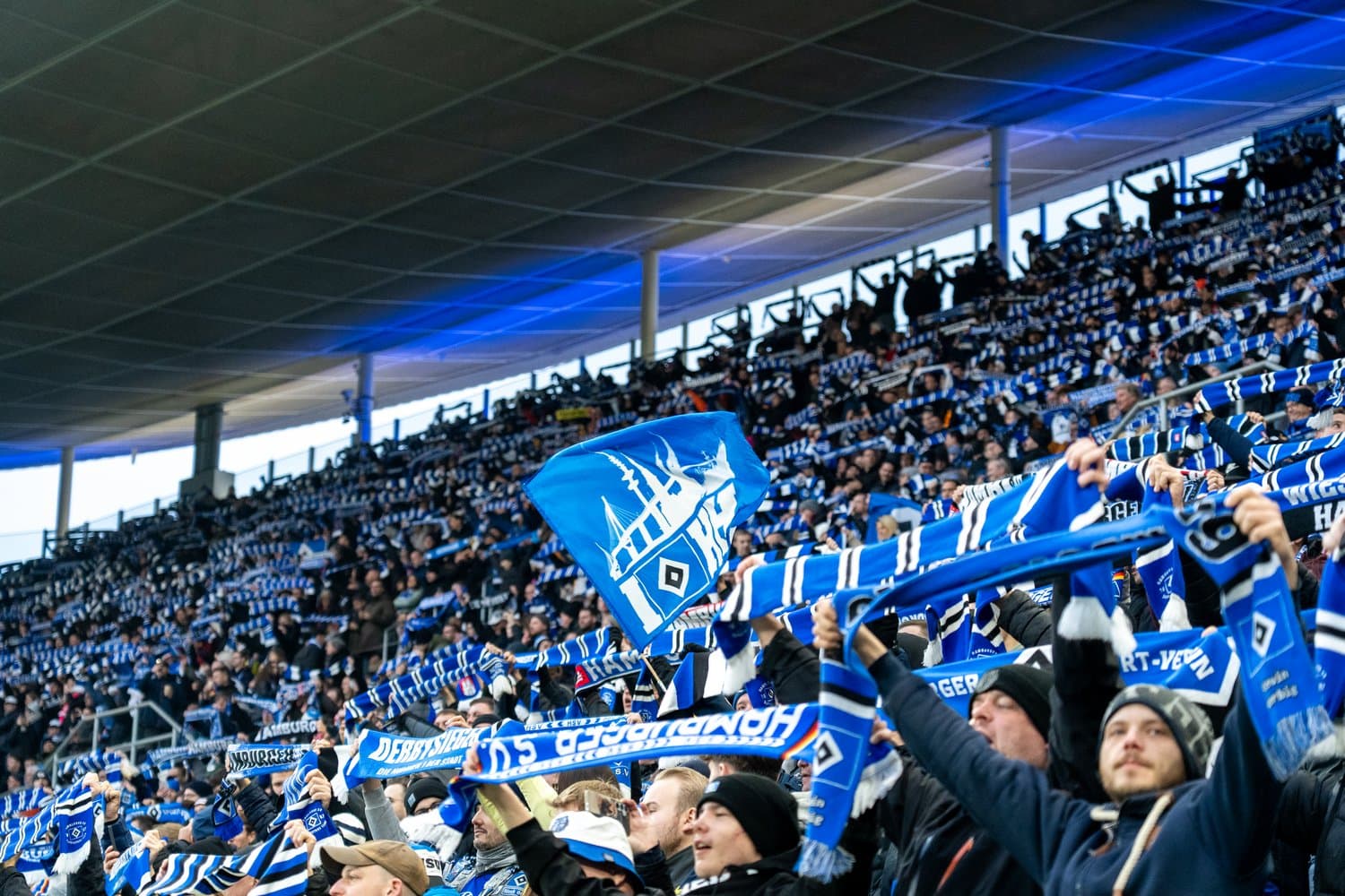 Fans in a stadium cheer energetically, waving blue and white scarves and flags, filling the stands with vibrant team colors.
